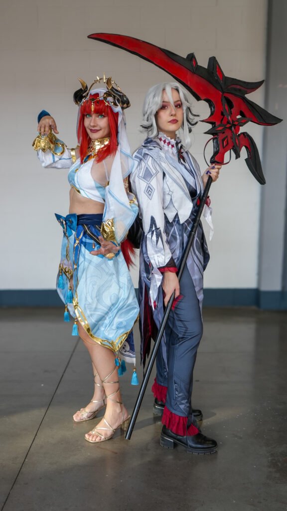 Due cosplayer in abiti fantasy, uno con capelli rossi e vestito azzurro e l'altro con capelli grigi e una falce rossa, posano in piedi durante un evento.