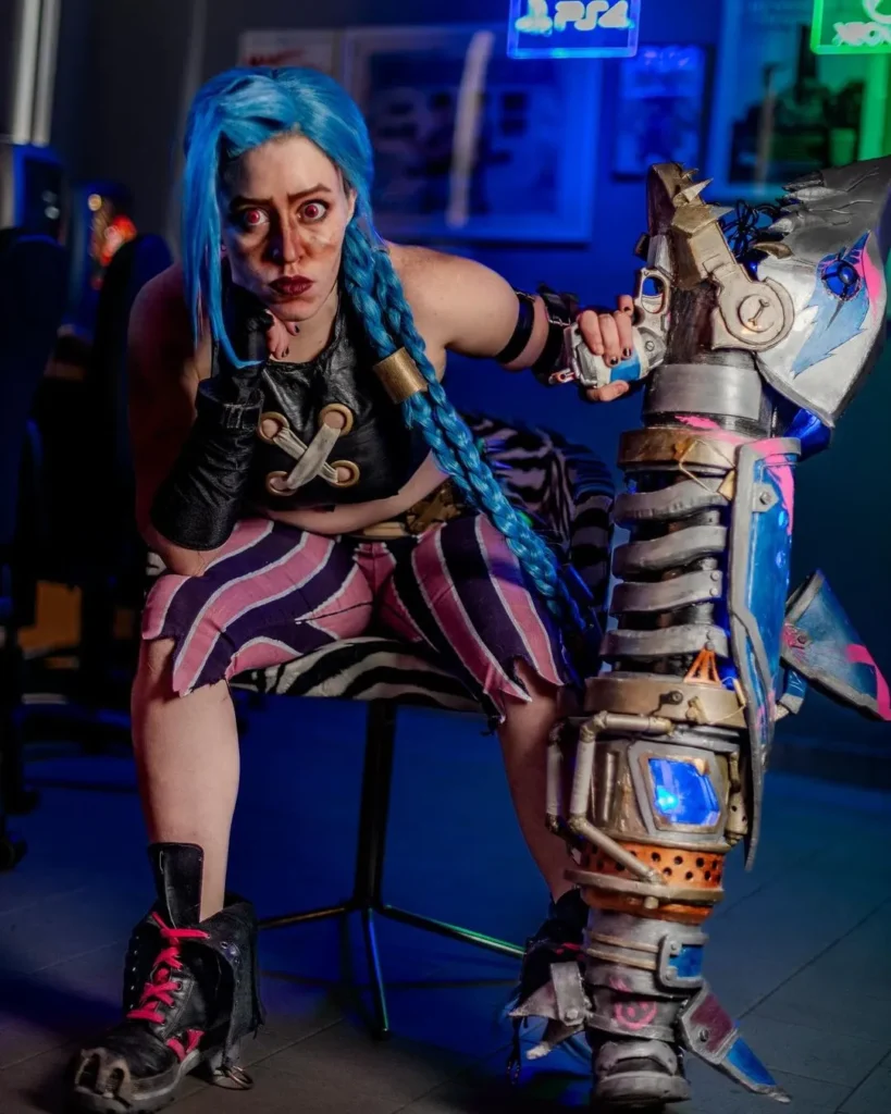 Lisanti Sara Cosplay Giudice Intervista Whimsi Community Cosplayer di Jinx da League of Legends con parrucca blu, seduta con espressione seria, in un'ambientazione retroilluminata con insegne al neon di console di videogiochi. Indossa abiti fedeli al personaggio e tiene in mano una grande arma ricostruita con dettagli luminosi