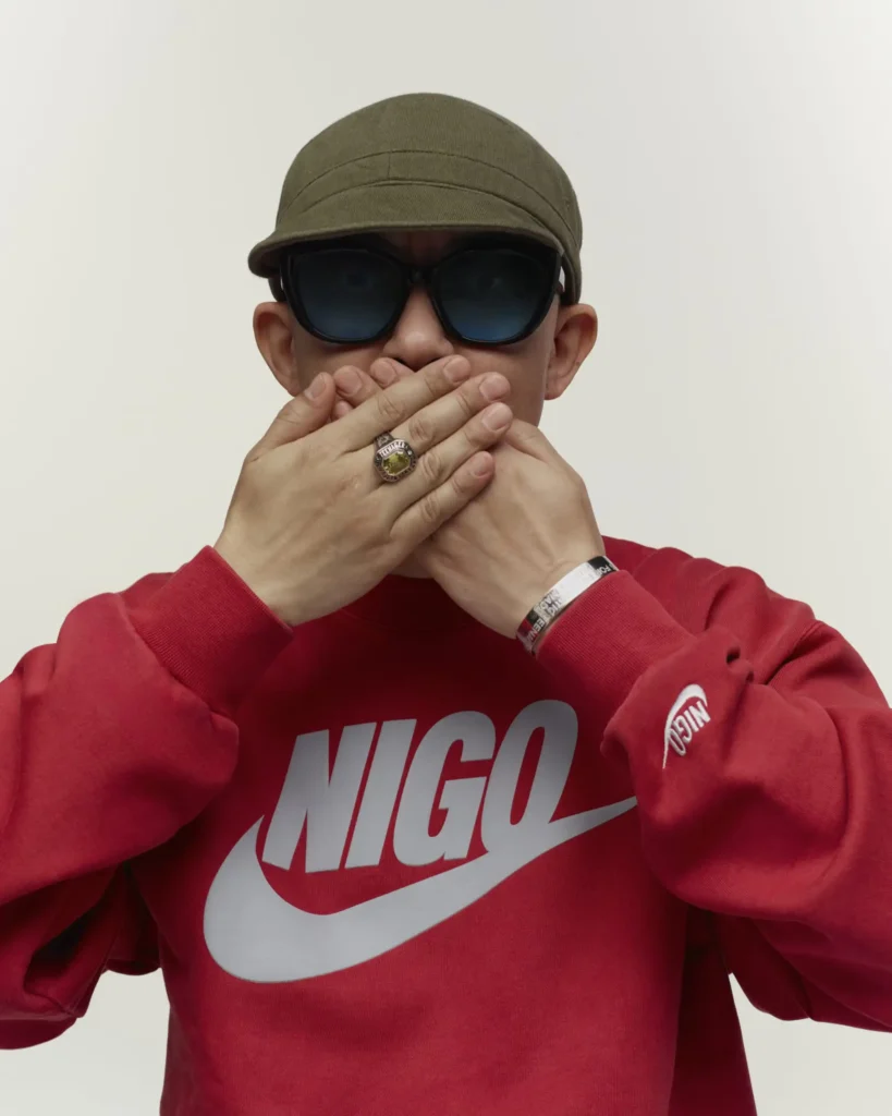 Nigo indossa una felpa Nike rossa con il suo nome e il logo Nike, coprendosi la bocca con le mani, occhiali da sole e cappello verde militare.