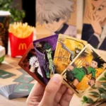 Una mano tiene quattro carte da collezione di Hunter x Hunter (Killua, Gon, Hisoka, e Chrollo), con il logo di McDonald's sullo sfondo, insieme a patatine fritte e un bicchiere decorato.