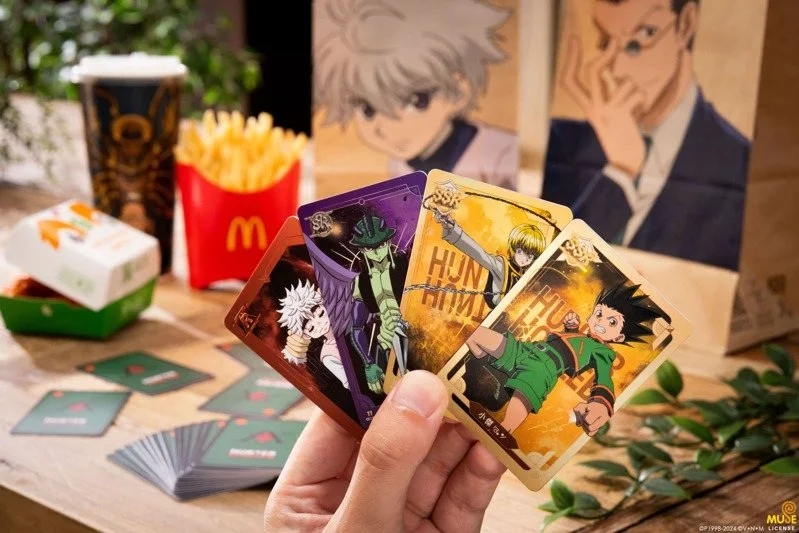 Una mano tiene quattro carte da collezione di Hunter x Hunter (Killua, Gon, Hisoka, e Chrollo), con il logo di McDonald's sullo sfondo, insieme a patatine fritte e un bicchiere decorato.